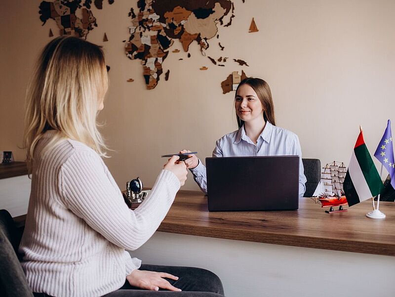 Eine Mitarbeiterin in einem Reisebüro sitzt lächelnd hinter einem Schreibtisch mit Laptop und kleinen Länderflaggen. Sie reicht einer Kundin, die ihr gegenüber sitzt, Reisedokumente oder Tickets. Im Hintergrund hängt eine dekorative Weltkarte an der Wand.