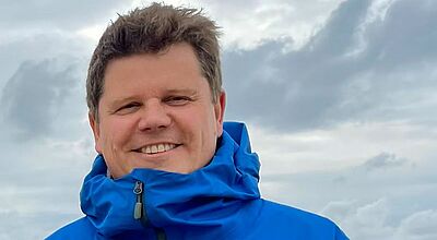 Ein Mann mittleren Alters mit braunem Haar steht in einen blauen Parka gehüllt vor weißen Wolken.