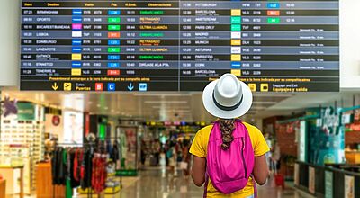 Reisebüros haben im vergangenen Touristikjahr viele Kunden in den Urlaub geschickt. Foto: Rafa Jodar/iStock Eine Person mit weißem Hut, gelbem T-Shirt und pinkem Rucksack steht in einem Flughafen-Terminal und blickt auf eine große Anzeigetafel mit Abflügen. Im Hintergrund sind Geschäfte und Reisende zu sehen.
