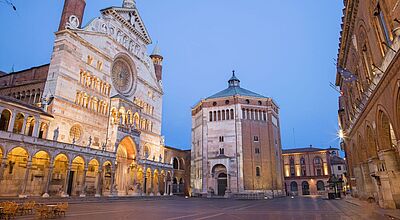 Eine der neuen Städtereisen von Avanti Busreisen hat die italienische Stadt Cremona zum Ziel. Foto: sedmak/istock Abendliche Ansicht des Domplatzes von Cremona in Italien mit der beleuchteten Fassade des Cremonaer Doms links, dem achteckigen Baptisterium in der Mitte und historischen Backsteingebäuden rechts, unter einem klaren blau-violetten Himmel.