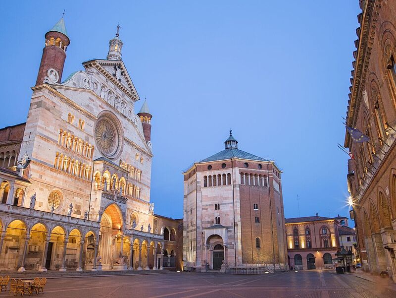 Eine der neuen Städtereisen von Avanti Busreisen hat die italienische Stadt Cremona zum Ziel. Foto: sedmak/istock Abendliche Ansicht des Domplatzes von Cremona in Italien mit der beleuchteten Fassade des Cremonaer Doms links, dem achteckigen Baptisterium in der Mitte und historischen Backsteingebäuden rechts, unter einem klaren blau-violetten Himmel.