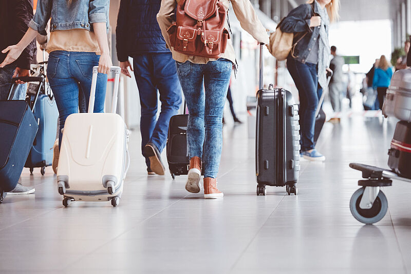Ansicht von hinten auf Reisende, die mit Rollkoffern und Taschen in einem belebten Flughafen-Terminal gehen.
