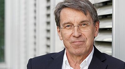 Porträt von einem Mann mittleren Alters, der lächelnd in die Kamera blickt. Er trägt ein weißes Hemd und ein dunkelblaues Sakko.