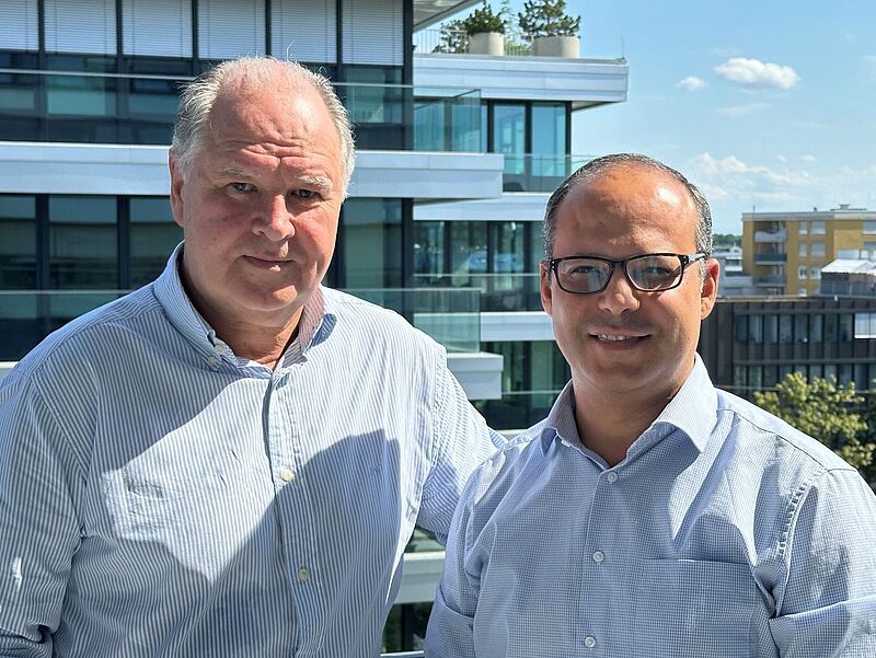 Zwei Männer in hellblauen Hemden stehen nebeneinander auf einer sonnigen Terrasse vor einem modernen Bürogebäude und lächeln in die Kamera.
