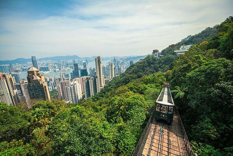 Atemberaubender Blick auf die Skyline von Hongkong mit dem Victoria Peak Tram, das durch üppige grüne Wälder den Berg hinauffährt – Panorama über den Victoria Harbour und die Hochhäuser der Stadt bei klarem Himmel.“