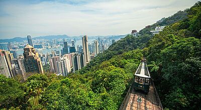 Modern und aufregend, aber auch grün: Hongkong ist das Zugpferd der Greater Bay Area Atemberaubender Blick auf die Skyline von Hongkong mit dem Victoria Peak Tram, das durch üppige grüne Wälder den Berg hinauffährt – Panorama über den Victoria Harbour und die Hochhäuser der Stadt bei klarem Himmel.“