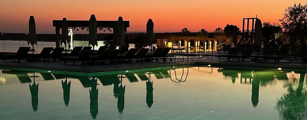 Ein ruhiger Außenpool bei Sonnenuntergang; das Wasser spiegelt die dunklen Umrisse von Liegen, geschlossenen Sonnenschirmen und einigen Bäumen. Am Horizont leuchtet ein sanfter Farbverlauf von Orange zu Dunkelblau, während rund um den Pool vereinzelte Lichter eine stimmungsvolle Atmosphäre erzeugen.