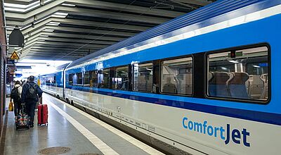 Ein moderner Fernverkehrszug der Tschechischen Bahn mit der Aufschrift „ComfortJet“ steht an einem überdachten Bahnsteig. Einige Reisende mit Gepäck laufen am Zug entlang in Richtung Einstieg. Durch die Fenster sind leere Sitzplätze im Inneren des Waggons sichtbar. Die Szene spielt sich an einem Bahnhof ab, der durch seine markante Deckenstruktur auffällt.