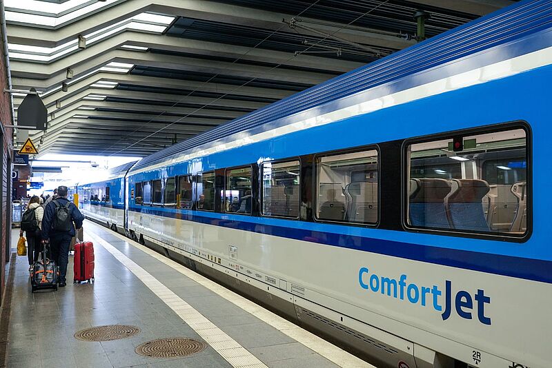 Ein moderner Fernverkehrszug der Tschechischen Bahn mit der Aufschrift „ComfortJet“ steht an einem überdachten Bahnsteig. Einige Reisende mit Gepäck laufen am Zug entlang in Richtung Einstieg. Durch die Fenster sind leere Sitzplätze im Inneren des Waggons sichtbar. Die Szene spielt sich an einem Bahnhof ab, der durch seine markante Deckenstruktur auffällt.