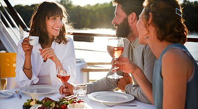 Auch der der Premium-alles-inklusive-Tarif wird in der Buchungsvorschau erläutert Zwei Frauen und ein Mann sitzen an einem gedeckten Tisch an Bord eines Flussschiffs und trinken Wein und Espresso