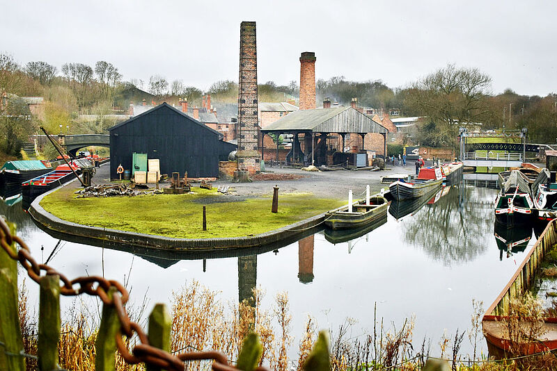 Historischer Industriekanal mit schwarzem Lagerhaus, hohen Backsteinschornsteinen, traditionellen Narrowboats und spiegelglattem Wasser