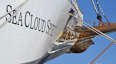 Am schneeweißen Bug eines modernen, luxuriösen Windjammers prangt der Schiffsname Sea Cloud Spirit. Taue sind vom Schiff an die Pier gespannt, im Hintergrund sind bunte Wohnhäuser auf einer Kanareninsel zu erkennen.