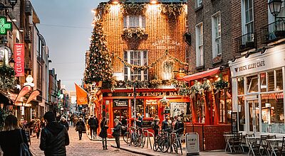 Zur Adventszeit erstrahl das berühmte Temple Bar Pub in der irischen Hauptstadt Dublin in einem besonderen Glanz. Bild einer belebten Straße zur Dunkelheit mit einem Backsteingebäude in dem sich eine beleuchtete Bar befindet.