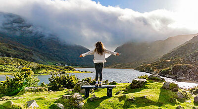 Eine Frau mit offenen Armen steht am See, umgeben vom Gebirge und genießt die Natur am Gap Of Dunloe im irischen County Kerry