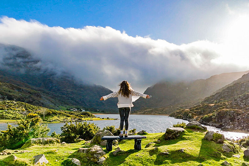 Eine Frau mit offenen Armen steht am See, umgeben vom Gebirge und genießt die Natur am Gap Of Dunloe im irischen County Kerry