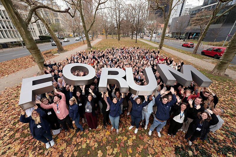 Rund 270 Reiseexperten diskutierten in Frankfurt am Main beim LCC-Forum über die Zukunft des Reisevertriebs Eine große Menschenmenge steht im Freien in einem herbstlichen Park und hebt gemeinsam ein großes, silberfarbenes 3D-Schriftobjekt mit dem Wort „FORUM“ in die Höhe. Die Personen lächeln, winken und wirken fröhlich; rundherum liegen gefallene Blätter, und im Hintergrund sind Bäume sowie angrenzende Straßen und Gebäude zu sehen.