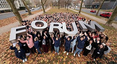 Eine große Menschenmenge steht im Freien in einem herbstlichen Park und hebt gemeinsam ein großes, silberfarbenes 3D-Schriftobjekt mit dem Wort „FORUM“ in die Höhe. Die Personen lächeln, winken und wirken fröhlich; rundherum liegen gefallene Blätter, und im Hintergrund sind Bäume sowie angrenzende Straßen und Gebäude zu sehen.