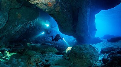 Zwei Taucher unter Wasser, die mit einem Licht ausgestattet durch eine unterirdische Höhle schwimmen.