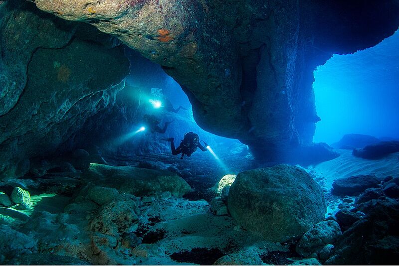 Rund um Malta können Höhlentaucher zum Beispiel Grotten oder Tunnel entdecken. Foto: Visit Malta Zwei Taucher unter Wasser, die mit einem Licht ausgestattet durch eine unterirdische Höhle schwimmen.