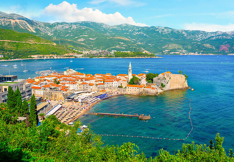 Mediterrane historische Stadt auf einer Landzunge umgeben von dunkelblauem Wasser, im Hintergrund ein grün bewachsenes Karstgebirge und blauem Himmel mit weißen Wolken. 