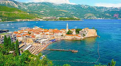 Mediterrane historische Stadt auf einer Landzunge umgeben von dunkelblauem Wasser, im Hintergrund ein grün bewachsenes Karstgebirge und blauem Himmel mit weißen Wolken. 