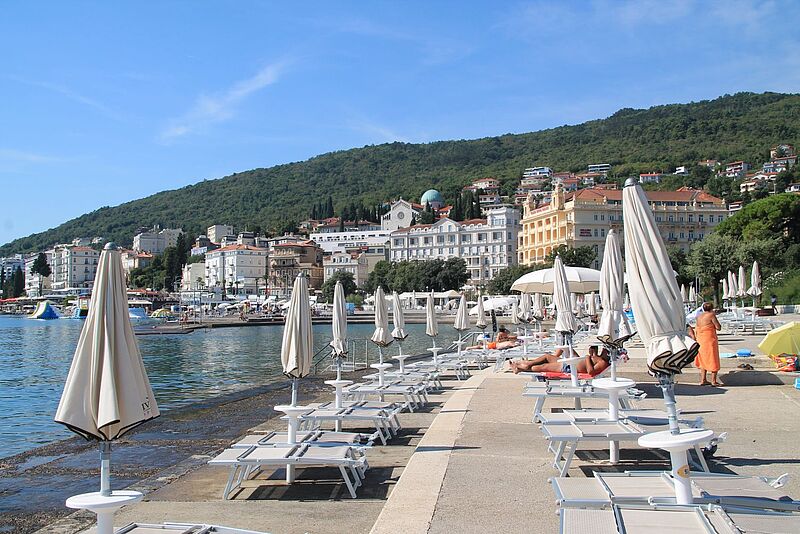 Eine steinerne Terrasse über dem Meer mit Liegen und Sonnenschirmen, Küste mit imposanten Gebäuden im Hintergrund