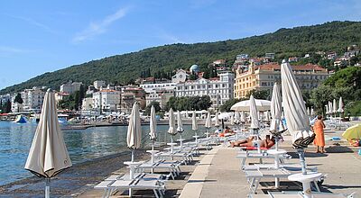 Eine neue Busreise nach Slowenien, Istrien und Triest führt auch ins Seebad Opatija. Eine steinerne Terrasse über dem Meer mit Liegen und Sonnenschirmen, Küste mit imposanten Gebäuden im Hintergrund