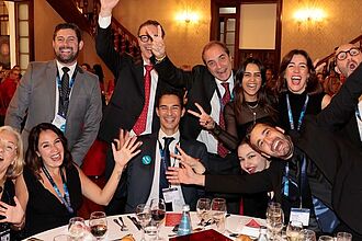 ine fröhliche Gruppe von Teilnehmerinnen und Teilnehmern sitzt und steht um einen festlich gedeckten Tisch in einem prunkvollen Saal. Alle lachen, winken und posieren ausgelassen in die Kamera während eines Abendessens bei der Lufthansa City Center Vollversammlung.