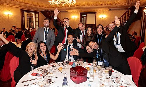 ine fröhliche Gruppe von Teilnehmerinnen und Teilnehmern sitzt und steht um einen festlich gedeckten Tisch in einem prunkvollen Saal. Alle lachen, winken und posieren ausgelassen in die Kamera während eines Abendessens bei der Lufthansa City Center Vollversammlung.