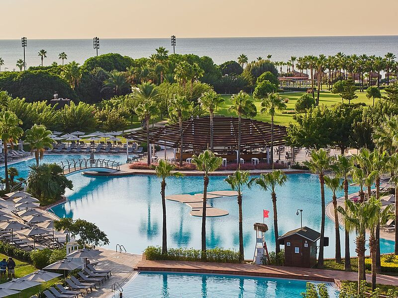 Ein luxuriöses Resort mit einer großen, geschwungenen Poollandschaft, umgeben von hohen Palmen und zahlreichen Sonnenliegen mit Sonnenschirmen. Eine Holzbrücke führt über den Pool, und in der Mitte des Wassers befindet sich eine Insel mit einer stilisierten Form. Im Hintergrund ist ein modernes, offenes Poolrestaurant mit einer überdachten Sitzfläche zu sehen. Dahinter erstreckt sich eine gepflegte Gartenanlage mit grünen Rasenflächen und weiteren Sitzbereichen, während das blaue Meer den Horizont bildet. Die Atmosphäre wirkt einladend und entspannend.