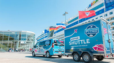 Blauer Shuttle-Van mit Anhänger vor Kreuzfahrtterminal, Logo „Parken & Meer Koffer Shuttle“ auf Fahrzeugen, Kreuzfahrtschiff im Hintergrund.