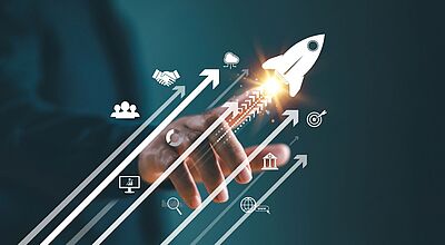 Im Vordergrund ist eine Hand zu sehen, die eine aufsteigende Rakete und Pfeile mit Geschäftssymbolen wie Zielscheibe, Netzwerk und Wachstum auslöst. Das Bild steht für Fortschritt, Start-up-Erfolg und Online-Business-Strategien. 