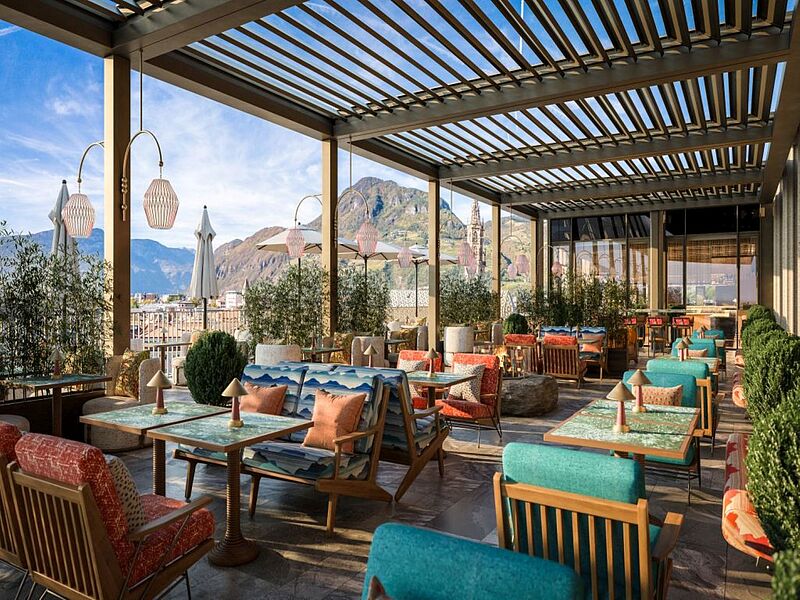 Tische mit bunten Stühlen auf einer Terrasse, im Hintergrund sind Berge zu erkennen