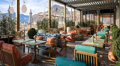 Tische mit bunten Stühlen auf einer Terrasse, im Hintergrund sind Berge zu erkennen