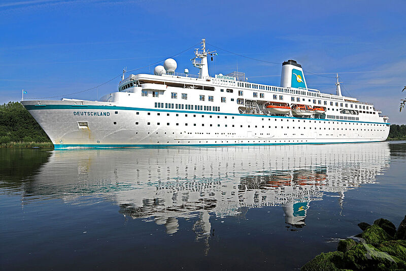 Seitliche Aufnahme des klassischen Kreuzfahrtschiffes Deutschland. Sein weißer Rumpf spiegelt sich auf der Wasseroberfläche.