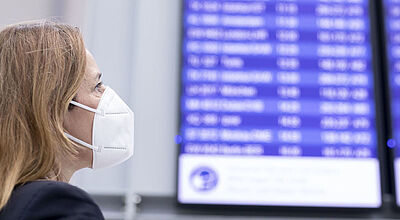 Eine Frau medizinischer Gesichtsmaske betrachtet auf einem Flughafen eine blaue Anzeigetafel mit Fluginformationen.