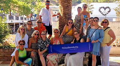 Gruppenfoto von 17 Personen, hauptsächlich Frauen, die unter einer großen Palme posieren. Die Gruppe gehört zur „Reiseberatung – powered by TUI“, was auf einem blauen Banner in der Mitte zu lesen ist. Im Hintergrund sind weiße Gebäude, Palmen und bewaldete Berge unter blauem Himmel zu sehen. Die Stimmung wirkt sonnig und entspannt – typisch für eine Reise in den Süden, vermutlich in Spanien.