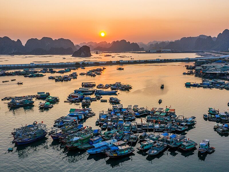 Luftaufnahme eines Fischerdorfs bei Sonnenuntergang mit zahlreichen bunten Fischerbooten, die dicht gedrängt im ruhigen Wasser liegen. Im Hintergrund erheben sich dunkle Kalksteinfelsen vor einem orangefarbenen Himmel, während die Sonne am Horizont über dem Meer untergeht. Rechts am Ufer sind Gebäude und ein kleiner Hafen zu erkennen.