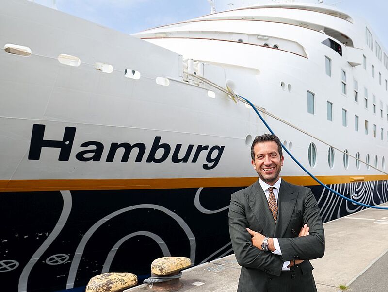 Inti Ligabue, Chef der Ligabue Group, will die Hamburg nach eigenen Plänen modernisieren lassen. Foto: Plantours Ein italienischer Reederei-Manager mit dunklen Haaren und in beigem Anzug steht lächelnd am Bug des an der Pier liegenden Kreuzfahrtschiffs Hamburg. Der Name des Schiffes prangt in großen Buchstaben an der Bordwand.