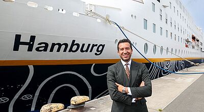 Ein italienischer Reederei-Manager mit dunklen Haaren und in beigem Anzug steht lächelnd am Bug des an der Pier liegenden Kreuzfahrtschiffs Hamburg. Der Name des Schiffes prangt in großen Buchstaben an der Bordwand.
