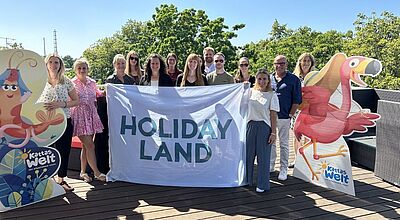Gruppenfoto von Teilnehmerinnen und Teilnehmern des Holiday-Land-Next-Generation-Events. Die Gruppe hält ein großes Banner mit der Aufschrift ‚Holiday Land‘. Links und rechts stehen bunte Pappfiguren aus ‚Katta’s Welt‘, darunter ein rotes Faultier und ein Vogel. Im Hintergrund sind Bäume und blauer Himmel zu sehen.