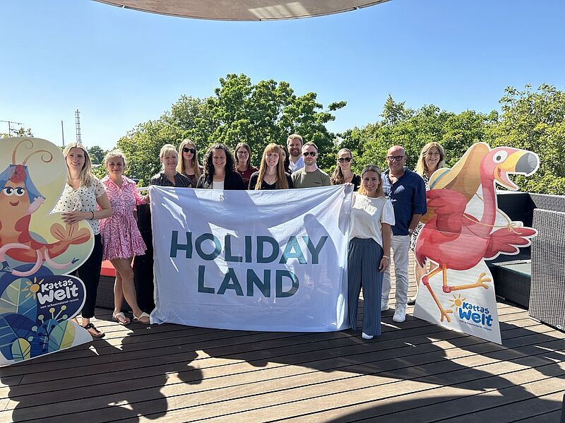 Gruppenfoto von Teilnehmerinnen und Teilnehmern des Holiday-Land-Next-Generation-Events. Die Gruppe hält ein großes Banner mit der Aufschrift ‚Holiday Land‘. Links und rechts stehen bunte Pappfiguren aus ‚Katta’s Welt‘, darunter ein rotes Faultier und ein Vogel. Im Hintergrund sind Bäume und blauer Himmel zu sehen.