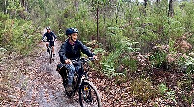 Zwei Personen fahren mit Fahrrädern auf einem schmalen Waldweg durch dichtes grünes Buschland. Sie tragen Helme und wetterfeste Kleidung, und der Weg ist mit Blättern bedeckt.
