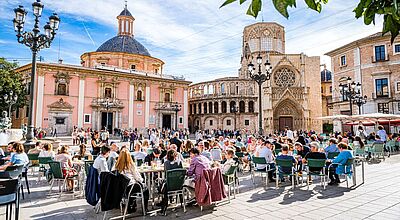 Viele Menschen sitzen in Restaurants auf einem Platz, umrundend von Kirchen, in der Frühlingssonne