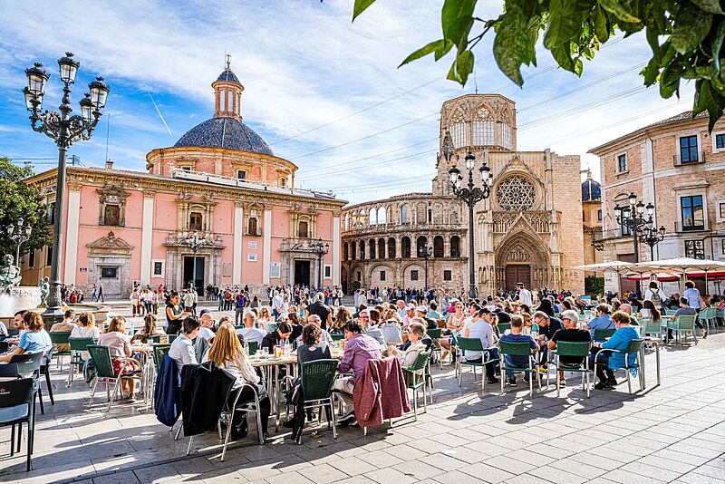 Viele Menschen sitzen in Restaurants auf einem Platz, umrundend von Kirchen, in der Frühlingssonne