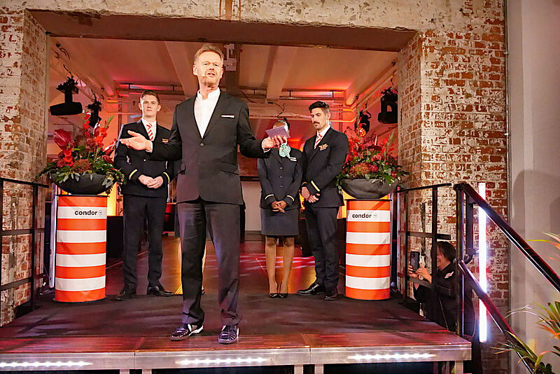 Condor-Chef Peter Gerber zog auf der Talk Night in Frankfurt eine positive Bilanz des Jahres Ein Manager mittleren Alters in einem dunklen Anzug steht gestikulierend auf einer bühnenartigen Treppe. Hinter ihm stehen unter einem backsteingemauerten Durchgang Airline-Mitarbeiter in Uniform sowie rot-weiß gestreifte Säulen mit der Aufschrift „Condor“, auf denen Blumensträuße stehen.