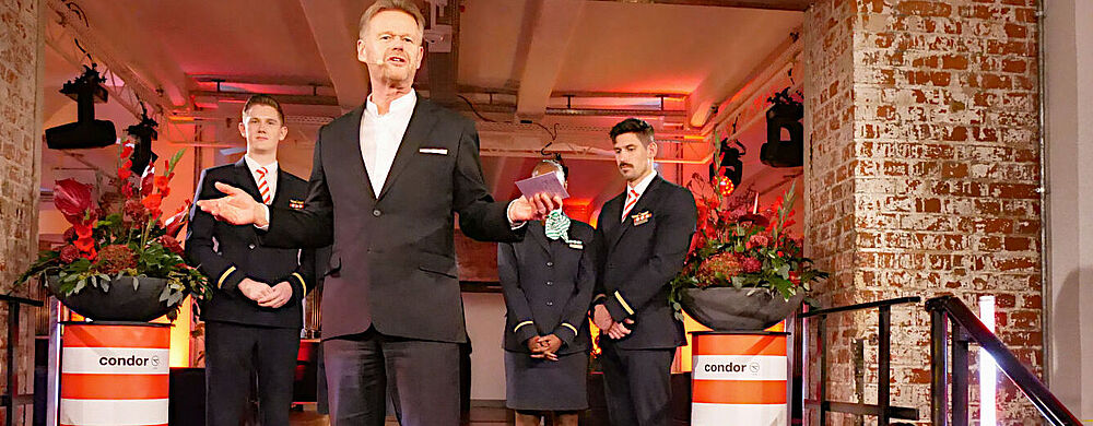 Condor-Chef Peter Gerber zog auf der Talk Night in Frankfurt eine positive Bilanz des Jahres Ein Manager mittleren Alters in einem dunklen Anzug steht gestikulierend auf einer bühnenartigen Treppe. Hinter ihm stehen unter einem backsteingemauerten Durchgang Airline-Mitarbeiter in Uniform sowie rot-weiß gestreifte Säulen mit der Aufschrift „Condor“, auf denen Blumensträuße stehen.