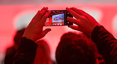 Mann fotografiert mit Handy Menschen auf einer rot beleuchteten Bühne