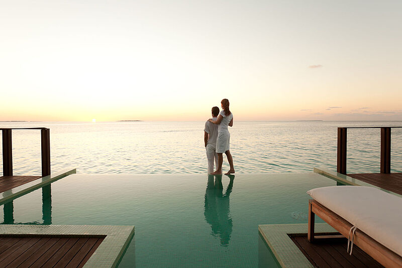 Ein junges Paar in weißer Sommerkleidung steht am Rand eines Infinity-Pools mit Blick auf den Sonnenuntergang und das Meer, umgeben von einer Holzterrasse und Liegemöbeln.