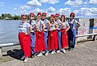 Eine Gruppe von acht Frauen posiert lachend am Ufer eines Flusses bei sonnigem Wetter. Sie tragen einheitliche Outfits bestehend aus weißen T-Shirts, roten Tüllröcken oder blauen Latzhosen, roten Mützen und Sonnenbrillen. Im Hintergrund ist Wasser, grüne Bäume und ein blauer Himmel mit weißen Wolken zu sehen.